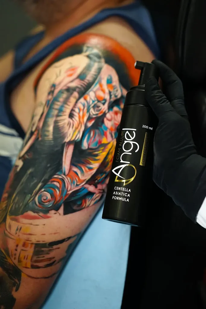 Gloved hand holding Angel Foam Allure tattoo aftercare foam cleanser next to a fresh, colorful tattoo.